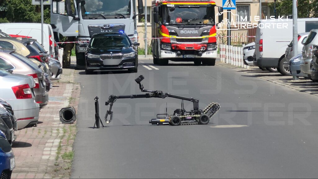 Robot z ramieniem przeszukującym jezdnię w otoczeniu zaparkowanych samochodów, obok policyjny radiowóz i wóz strażacki.