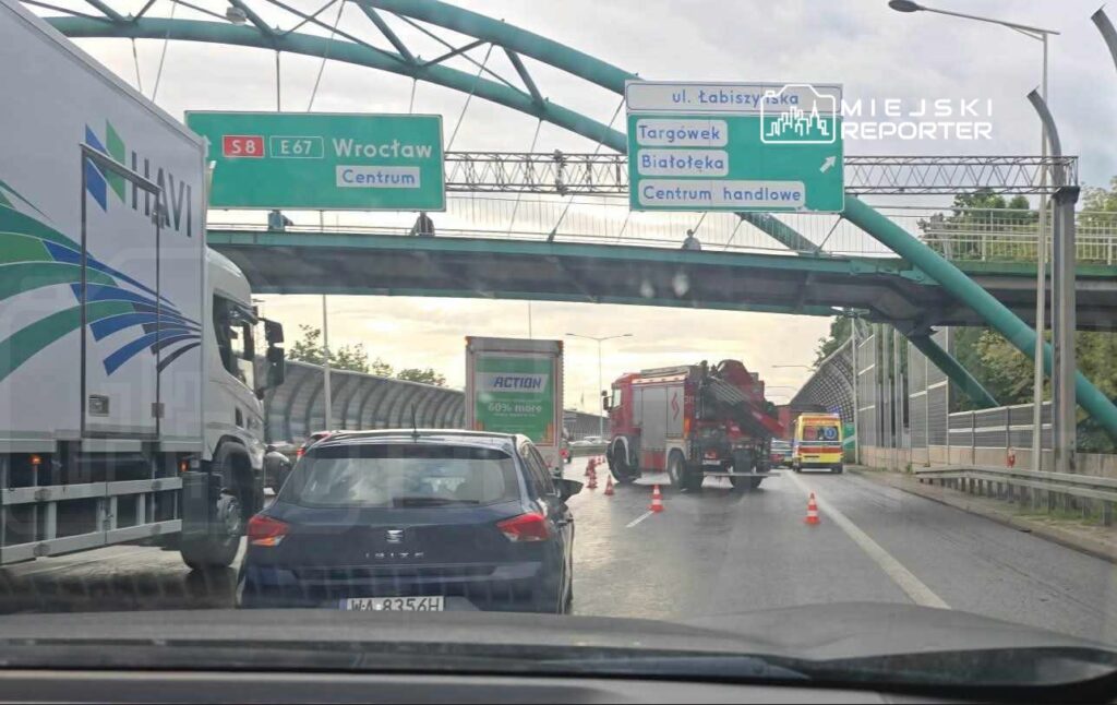 Ruch samochodowy wstrzymany na drodze, obok pojazdy ciężarowe oraz straż pożarna z włączonymi sygnałami.
