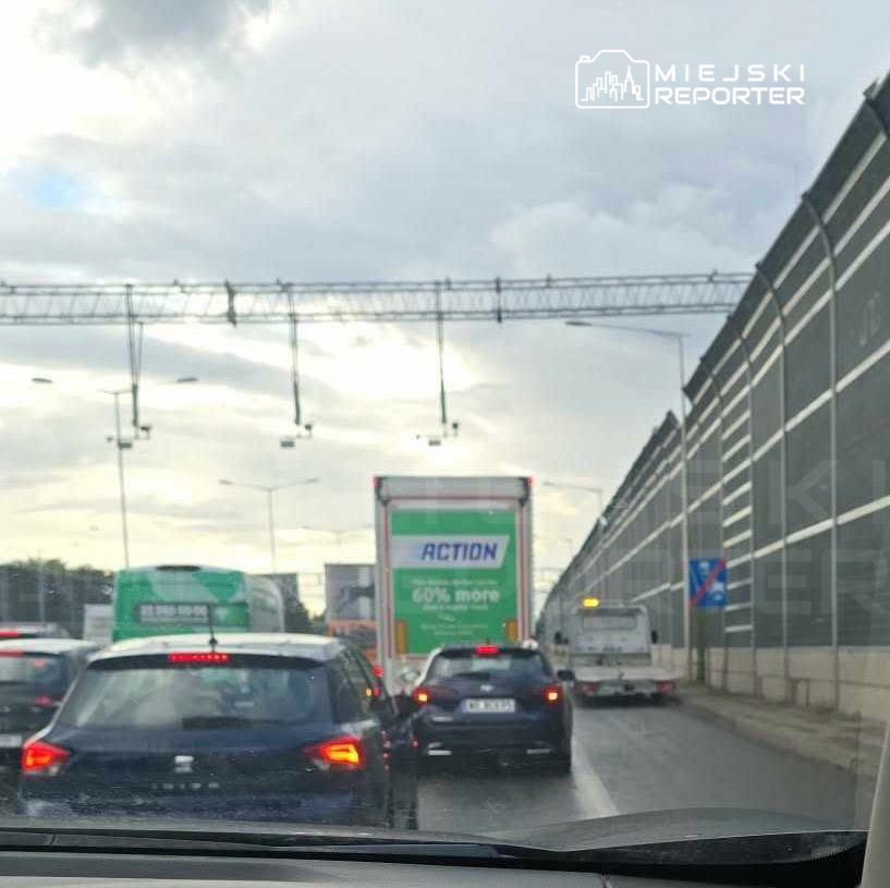 Kierowcy stoją w korku na ruchliwej drodze, mijając ciężarówkę z napisem "ACTION" oraz oznakowanie drogowe.
