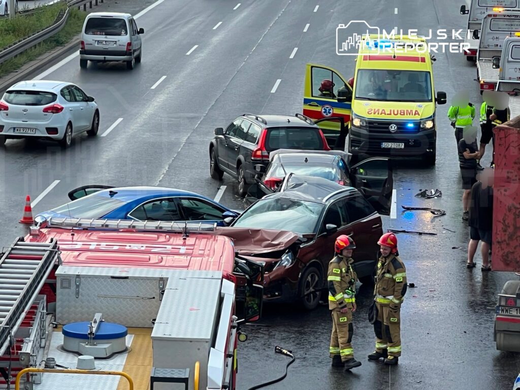 Wielokrotny wypadek samochodowy na drodze, z pojazdami zderzonymi, a obok strażacy rozmawiający przy wozie strażackim.