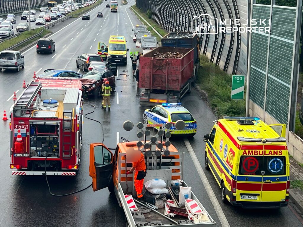 Na drodze widać wypadek samochodowy z uszkodzonymi pojazdami, w tym czerwonym autem. Służby ratunkowe udzielają pomocy.
