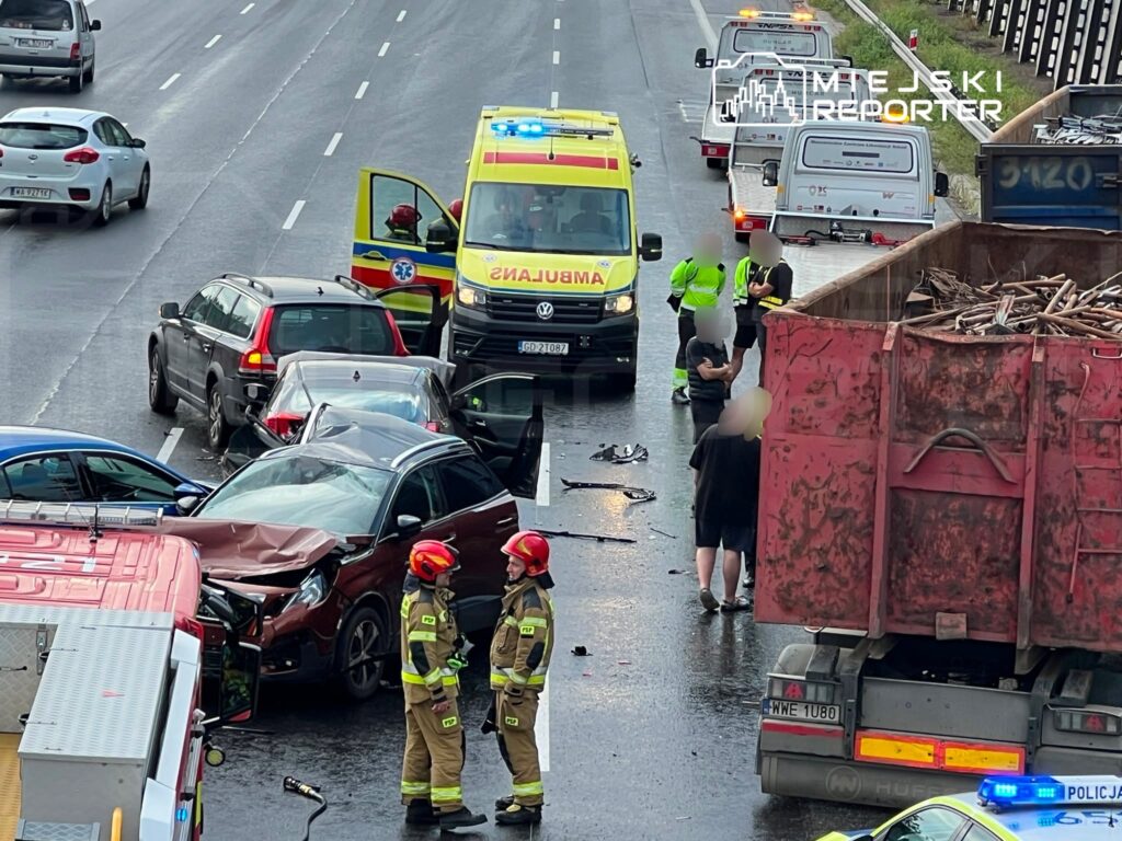Wielokrotny wypadek drogowy z udziałem dwóch rozbitych samochodów osobowych oraz ciężarówki na jezdni. Ratownicy medyczni udzielają pomocy poszkodowanym. W tle widać pojazdy służb ratunkowych oraz zablokowany ruch.