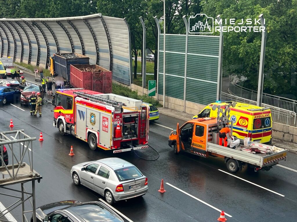 Na jezdni zatrzymane pojazdy, strażacy udzielają pomocy poszkodowanym, a obok pracownicy służby drogowej instalują znaki.