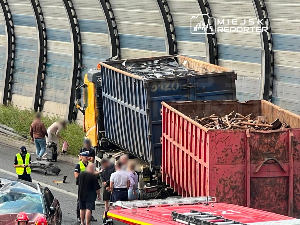 Na drodze stoi przewrócona ciężarówka z metalowymi odpadami, wokół zbiera się grupa ludzi oraz służby ratunkowe.