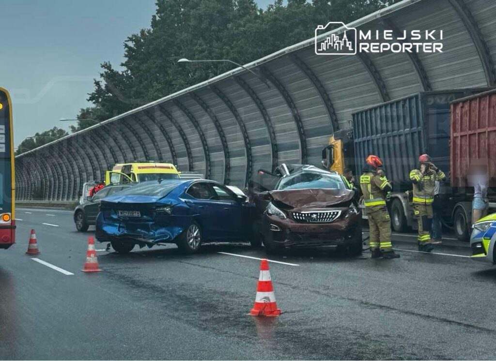 Dwa uszkodzone samochody osobowe na drodze, obok pracują strażacy w kamizelkach odblaskowych.