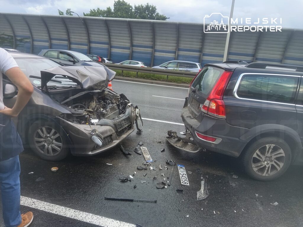 Zderzenie dwóch samochodów na drodze, jeden z pojazdów ma uszkodzoną przednią część, a drugi tylną. W tle widać inne samochody.