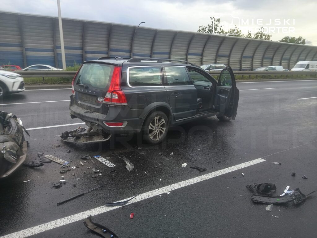 Uszkodzony samochód osobowy stoi na poboczu drogi, obok widać fragmenty zderzaka oraz rozbite elementy.