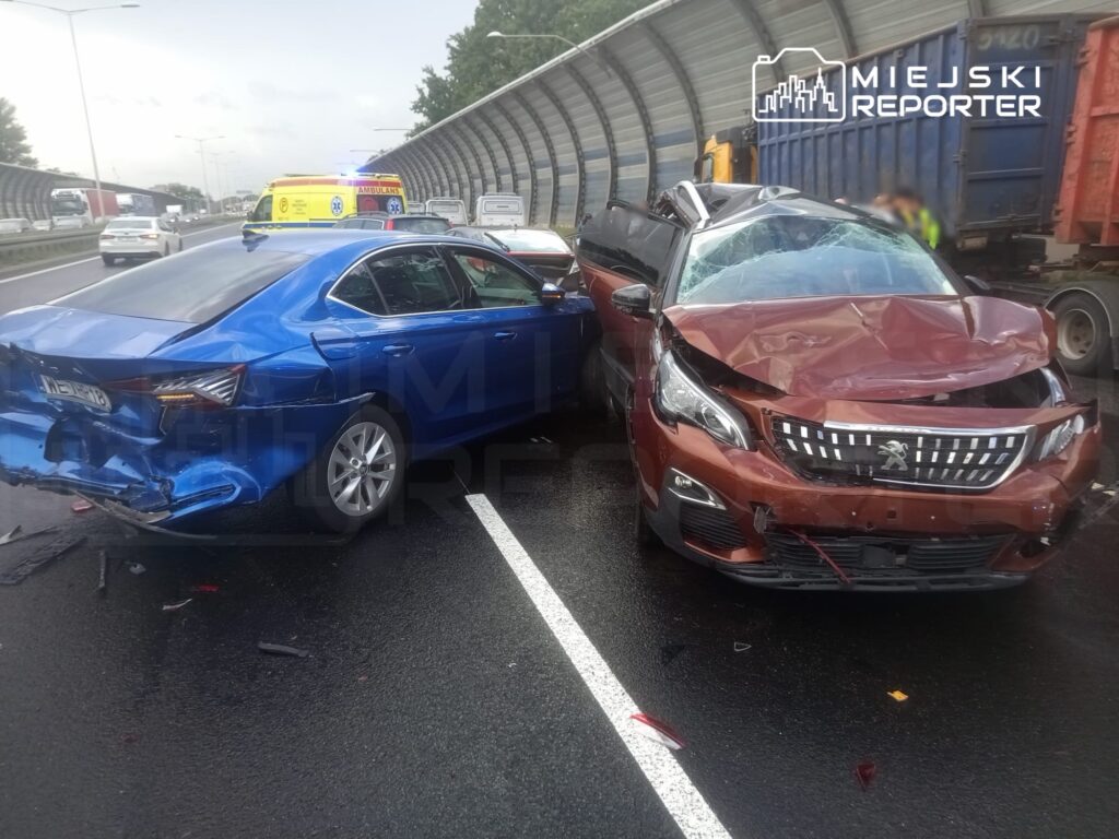 Zderzenie dwóch uszkodzonych samochodów osobowych na drodze, w tle widać ambulans oraz inne pojazdy.