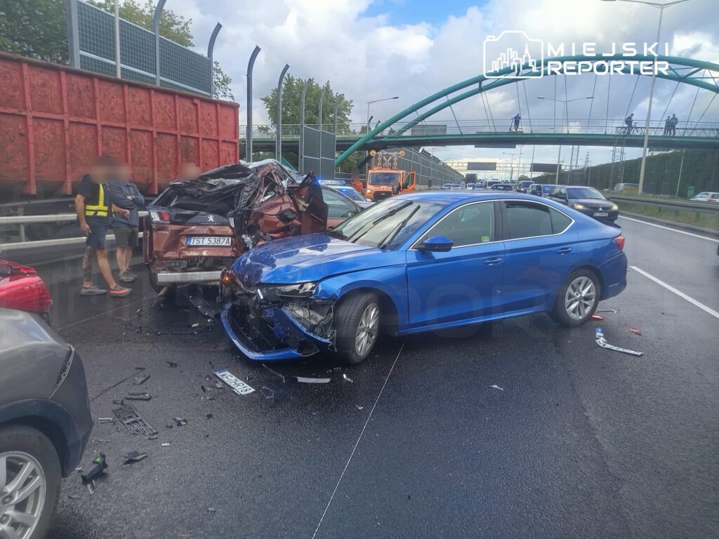 Zderzenie dwóch samochodów na drodze, z uszkodzonym niebieskim sedanem i brązowym pojazdem, w tle widać ciężarówkę.
