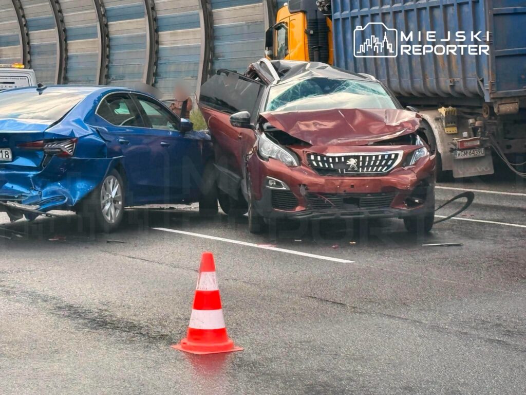 Dwa uszkodzone samochody osobowe na drodze, z czerwoną limuzyną z wgniecionym przodem i niebieską z uszkodzoną tylną częścią. W pobliżu stoi pachołek drogowy.