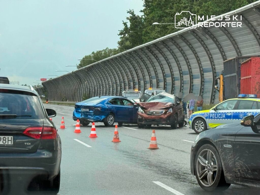Na skrzyżowaniu drogi stoją uszkodzone samochody, a obok nich policja zabezpiecza miejsce wypadku.