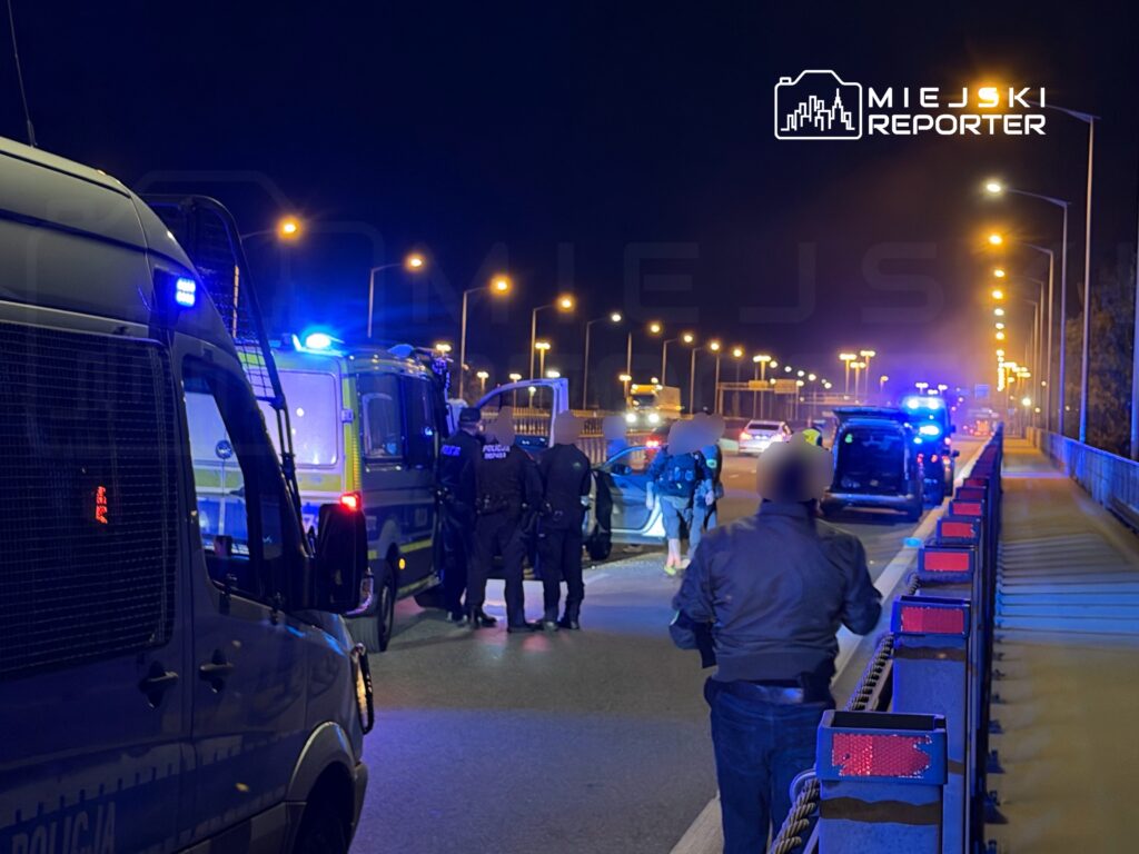Grupa policjantów w mundurach przeprowadza kontrolę drogową na oświetlonym moście w nocy, obok zaparkowanych radiowozów.