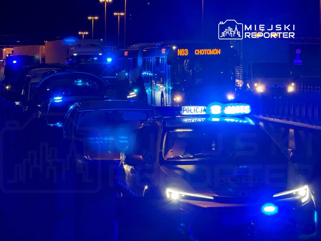Policyjny radiowóz z włączonymi sygnalizatorami świetlnymi stoi w zatorze drogowym, obok przystanku autobusowego.