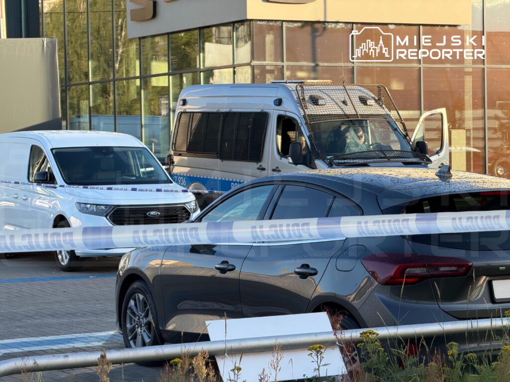 Policyjny radiowóz oraz biały furgon zaparkowane przy budynku, otoczone taśmą zabezpieczającą.