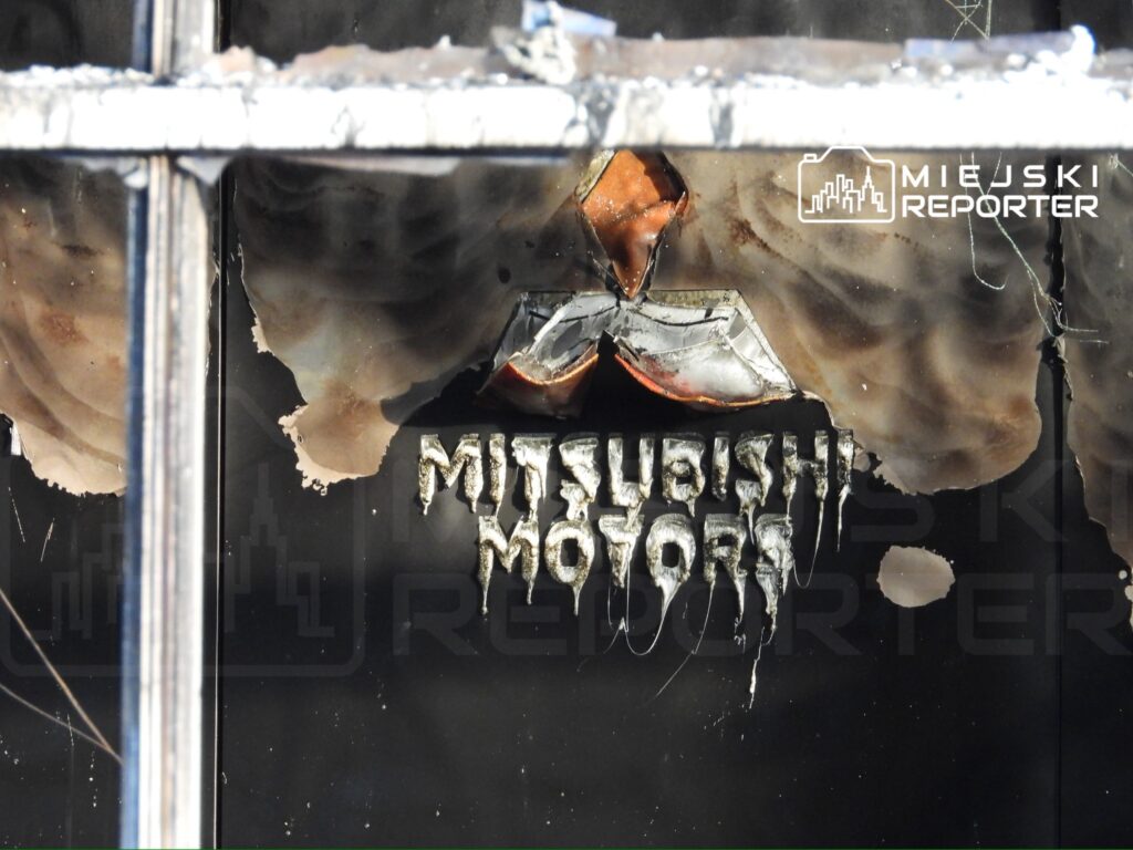 Uszkodzony szyld z napisem "Mitsubishi Motors" z widocznymi śladami po pożarze w zniszczonym budynku.
