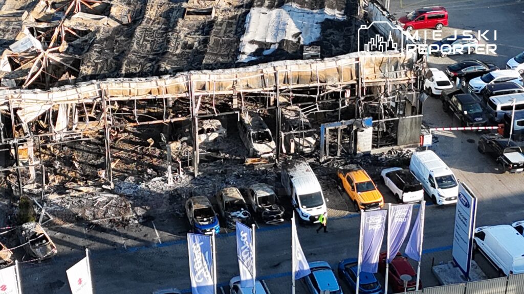 Zniszczony budynek z palącymi się fragmentami dachu oraz spalonymi pojazdami na parkingu obok.