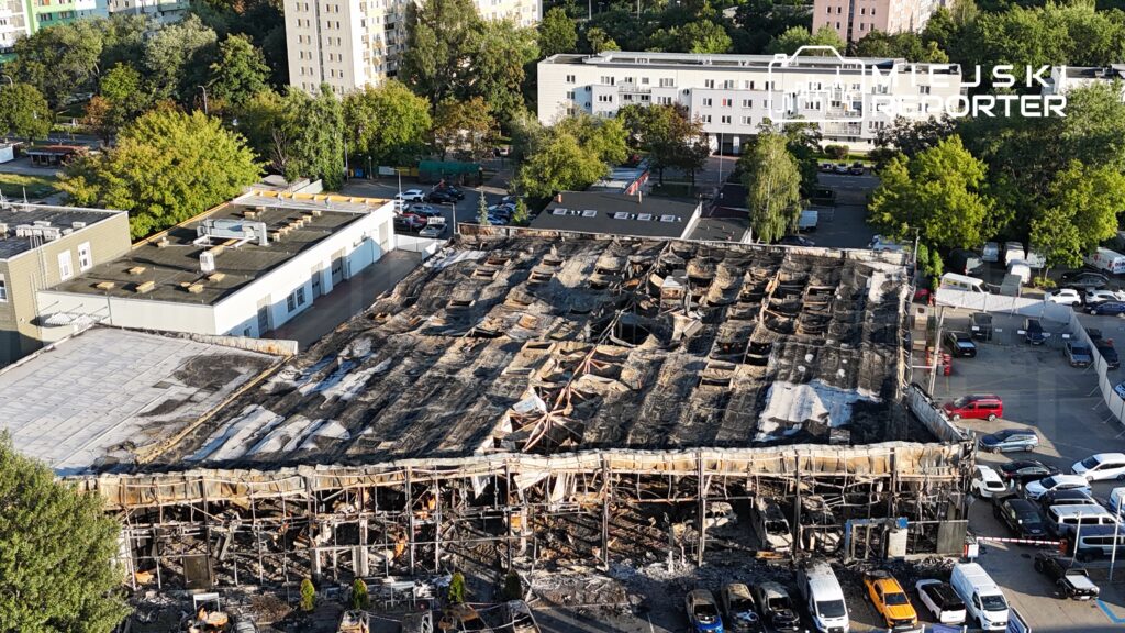 Zniszczony budynek z uszkodzoną dachówką i spalonymi elementami w otoczeniu zieleni i zaparkowanych samochodów.