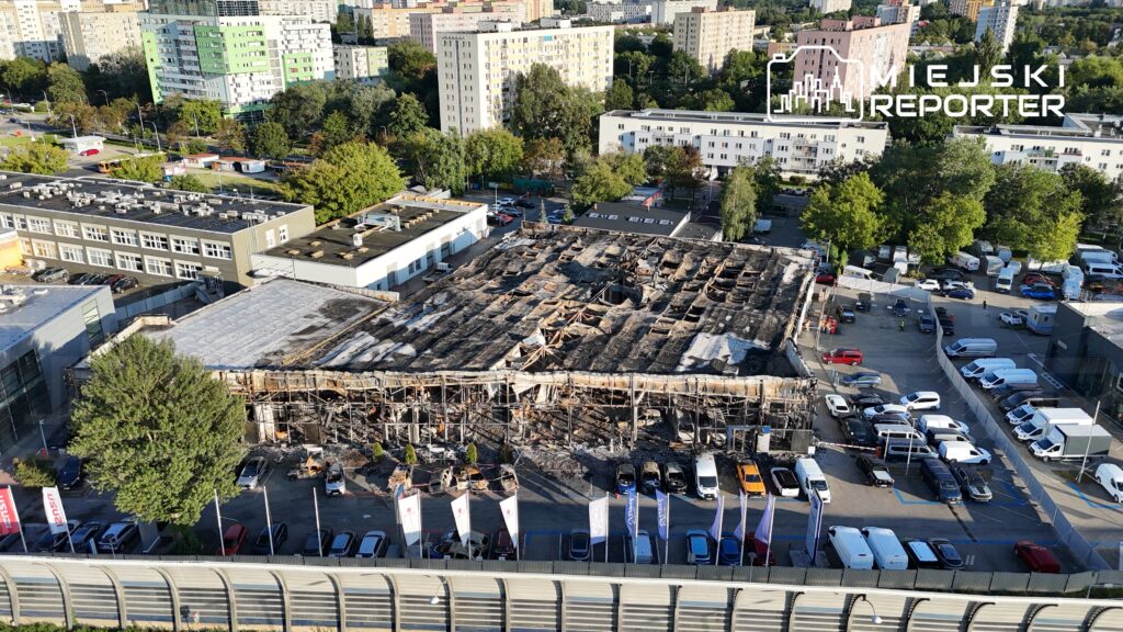 Zniszczony budynek z wypalonym dachem, otoczony parkingiem z samochodami oraz drzewami w tle.