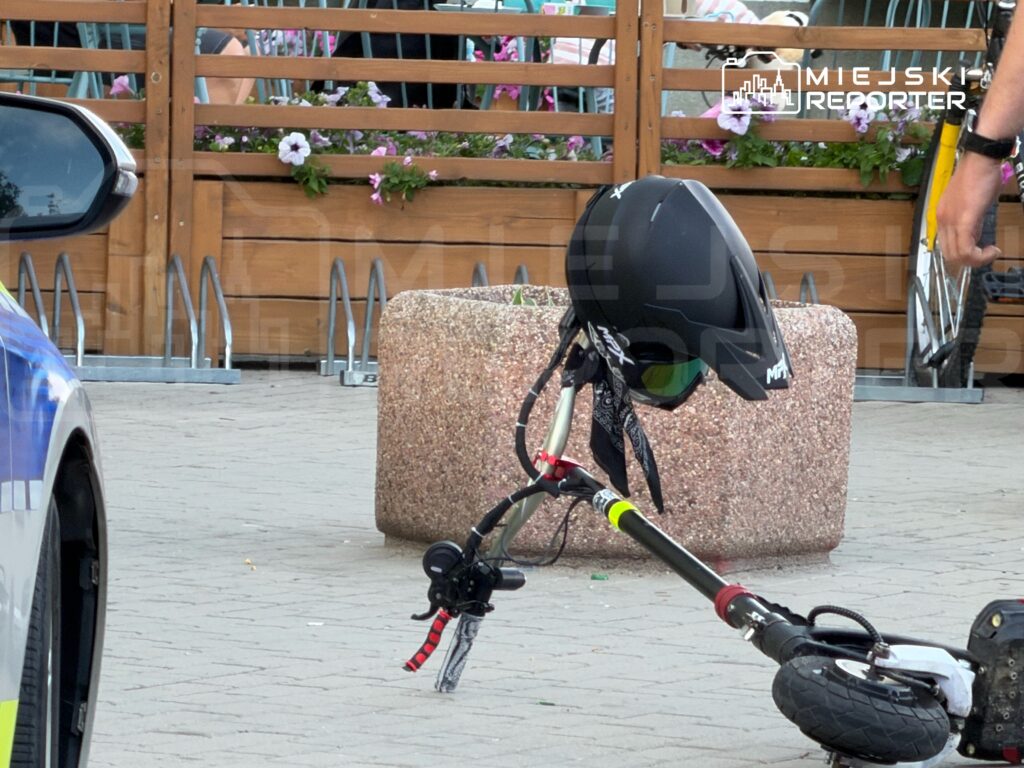 Skuter elektryczny z kaskiem leżący obok granitowego słupka w otoczeniu restauracji i rowerów.
