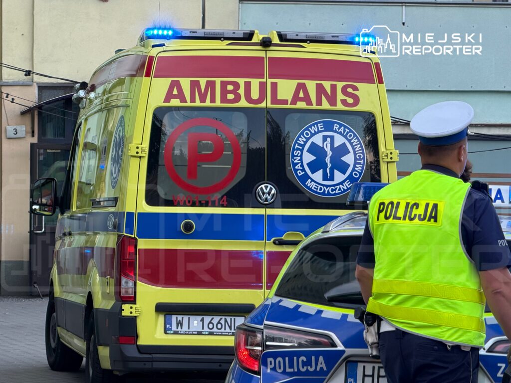 Ambulans z włączonymi sygnałami świetlnymi stoi na ulicy, obok policjant w kamizelce odblaskowej rozmawia z kolegą.