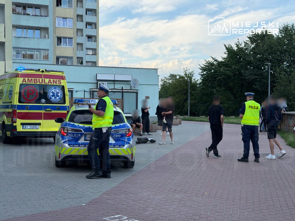 Grupa osób stoi na chodniku obok policyjnego radiowozu i karetki, w tle widoczny budynek mieszkalny.