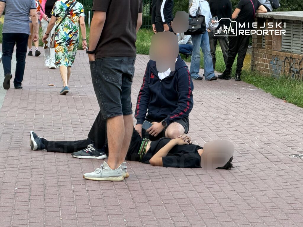 Mężczyzna siedzący na chodniku, trzymający leżącą osobę, podczas gdy przechodnie mijają ich obok.