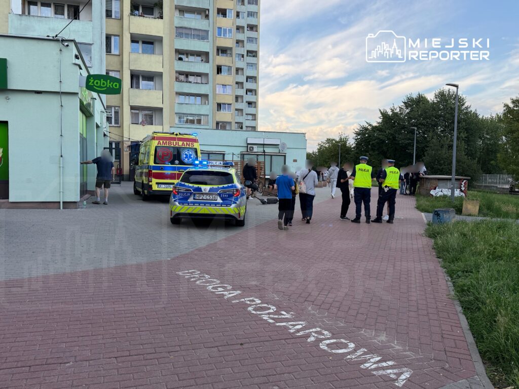 Grupa osób rozmawia z policjantami przy ambulansie w pobliżu sklepu Żabka, w tle bloki mieszkalne.