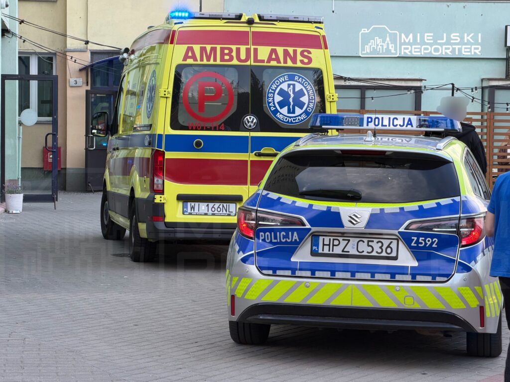 Ambulans z napisem "AMBULANS" oraz radiowóz policyjny zaparkowane przed budynkiem, w tle widoczny napis "MIEJSKI REPORTER".