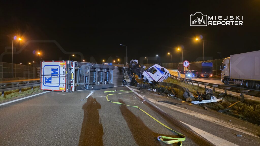 Przewrócony ciężarowy pojazd z uszkodzoną kabiną leży na drodze, obok widać uszkodzenia bariery ochronnej.