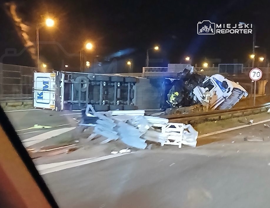 W nocy przewrócony ciężarówka leży na drodze, a obok pracują strażacy. W tle widoczne latarnie uliczne.