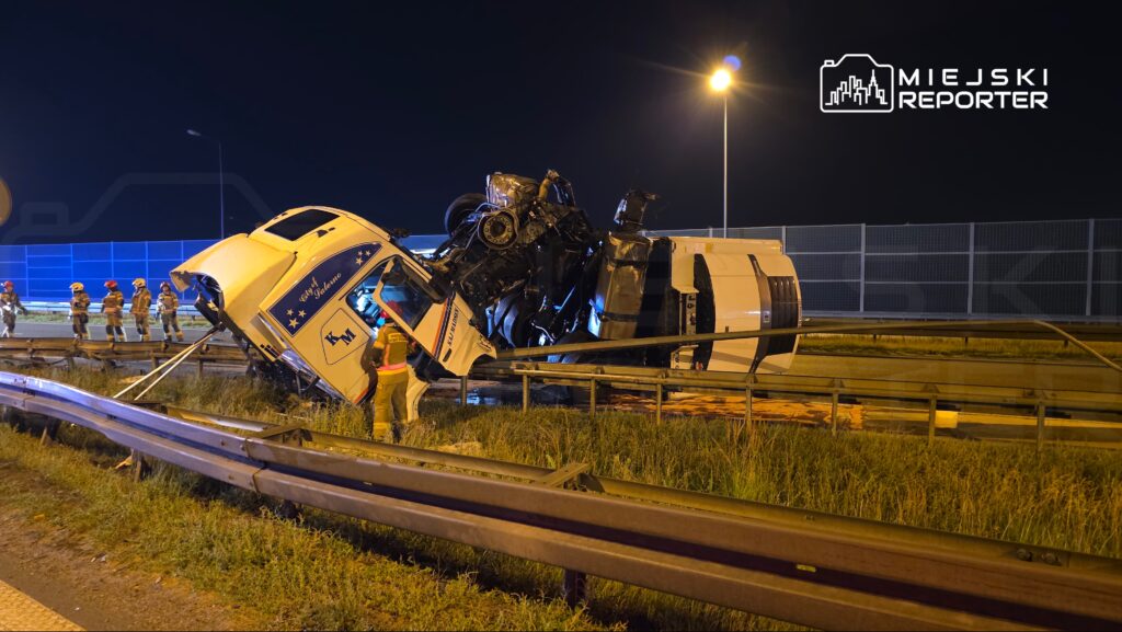 Cysterna przewrócona na drodze, obok grupka strażaków w hełmach, w tle nocne oświetlenie.