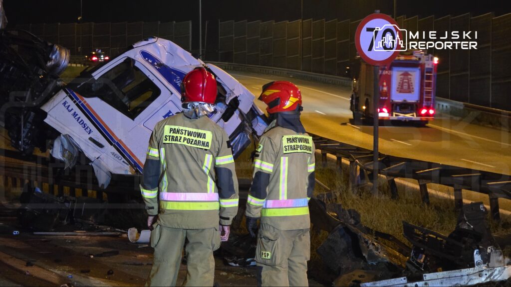 Dwaj strażacy w mundurach z odblaskowymi elementami obserwują przewrócony pojazd ciężarowy na autostradzie.