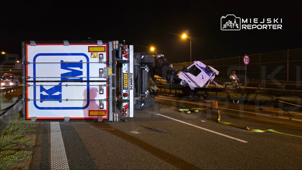 Przewrócony ciężarówka leży na drodze, a obok pracują ratownicy w odblaskowych kamizelkach.