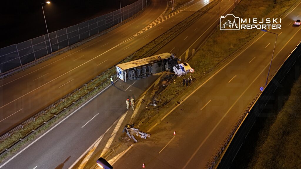 W nocy strażacy i policja pracują przy przewróconym autobusie na drodze, obok leży uszkodzony samochód.