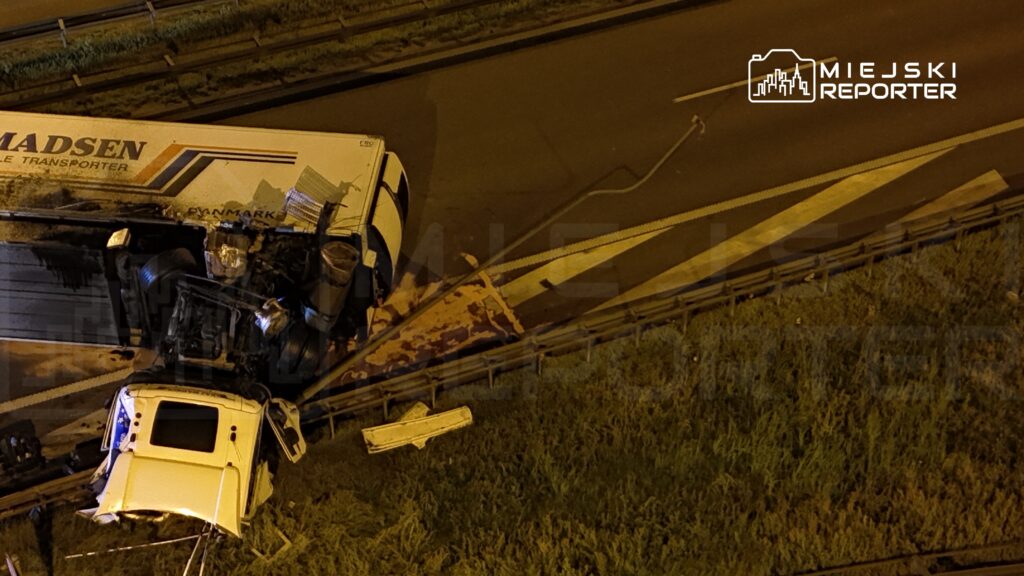 Wywrócony ciężarówka leży na poboczu drogi, z uszkodzoną kabiną i przewróconymi kołami. Obok widoczna jest zniszczona barierka ochronna.