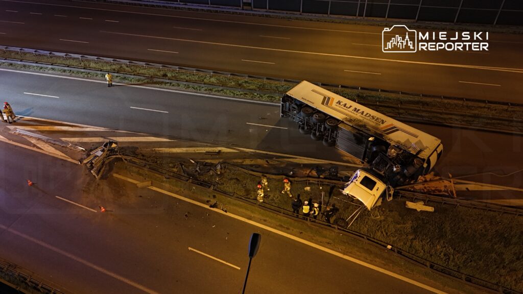 Na drodze leży przewrócony ciężarówka, a obok pracują strażacy w odblaskowych kamizelkach.