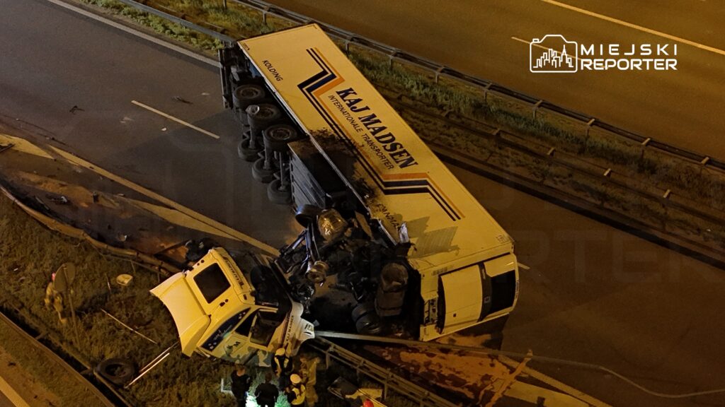 Ciężarówka leżąca na boku na pasie ruchu, obok stoją ratownicy oraz pojazdy służb ratunkowych.