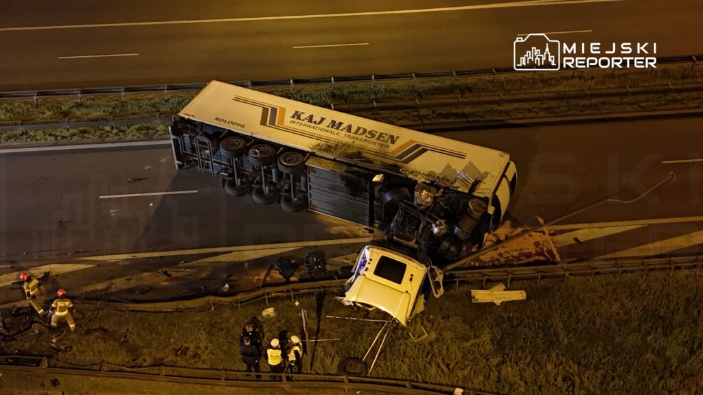 Czterech ratowników pracuje przy przewróconej ciężarówce na autostradzie, obok leży uszkodzony pojazd osobowy.