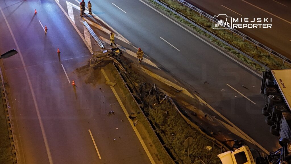 Grupa strażaków pracuje nad usunięciem zniszczonego bariery ochronnej na drodze, obok przewróconej ciężarówki.