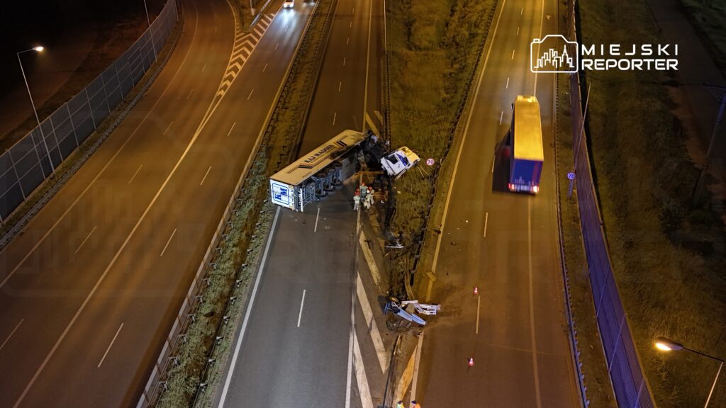 W nocy na drodze leży przewrócony tir, obok grupka ratowników. W tle widoczny inny ciężarówka i oświetlona jezdnia.