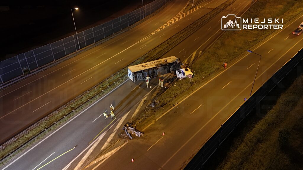Na drodze leży przewrócony autobus, a obok niego pracują ratownicy w odblaskowych ubraniach. W tle widoczne są samochody i latarnie.