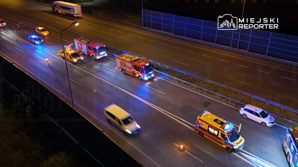Na drodze nocą znajdują się pojazdy służb ratunkowych, w tym dwa wozu strażackie, ambulans oraz policyjny, z włączonymi sygnalizacjami świetlnymi.