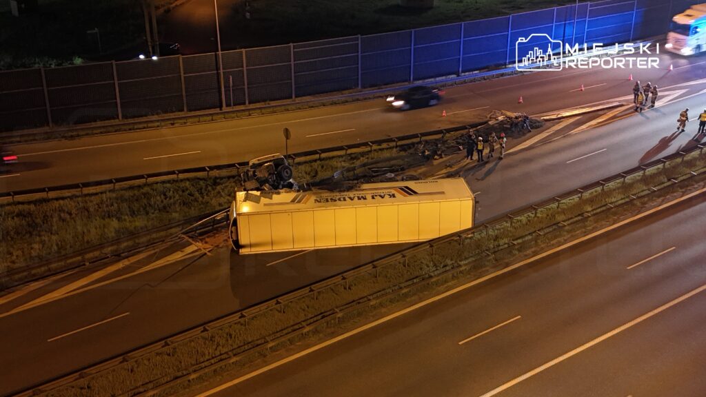 Na autostradzie leży przewrócony ciężarówka, wokół której stoją strażacy i policjanci, a w tle widać inne pojazdy.