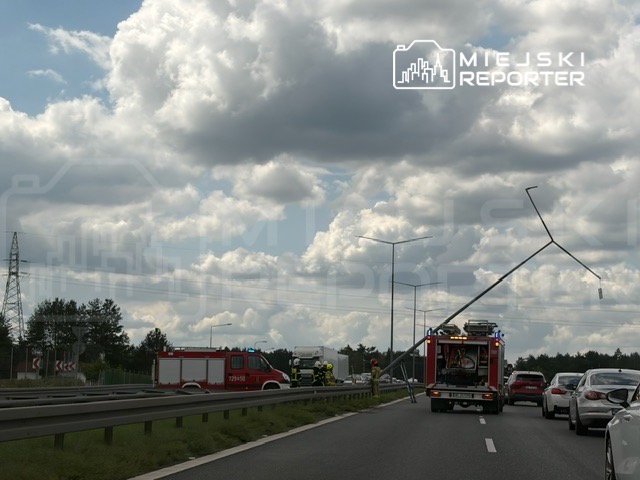 Strażacy pracują przy uszkodzonej latarni na drodze, obok stojące samochody i pojazd strażacki.