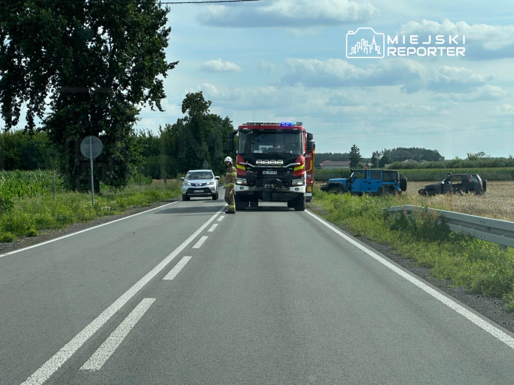 Straż pożarna zatrzymana na drodze, obok samochód osobowy, a w tle dwa pojazdy terenowe na poboczu.