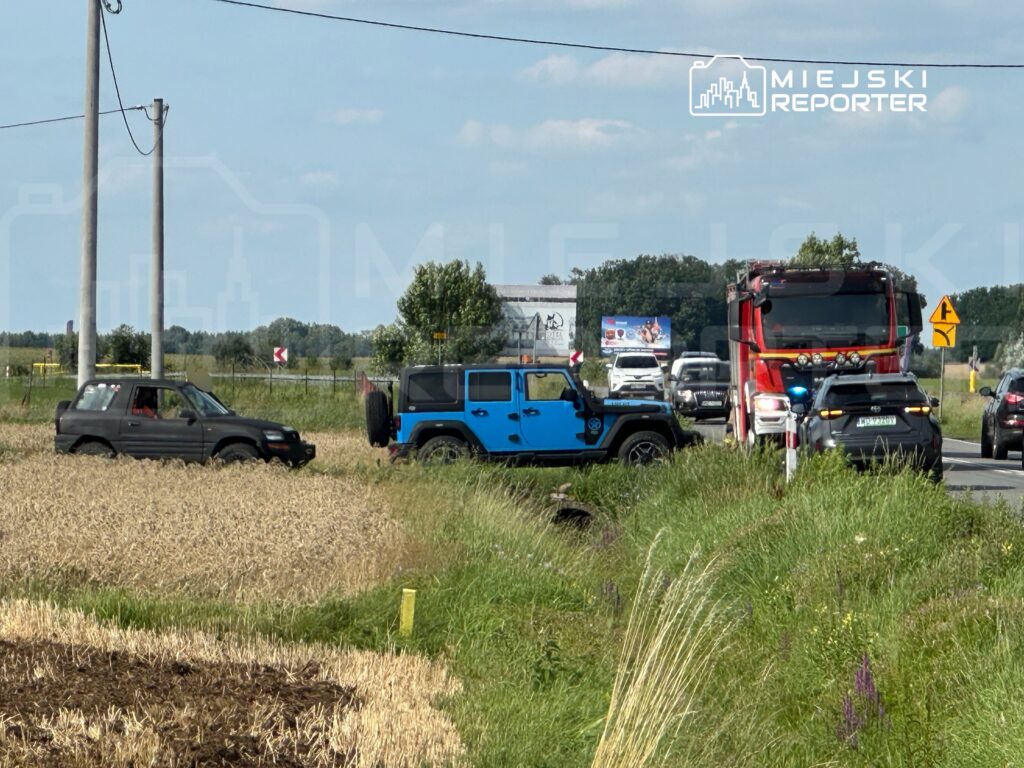 Na drodze widać dwa samochody osobowe, z których jeden utknął w polu, obok stoi nieoznakowany radiowóz oraz wóz strażacki.