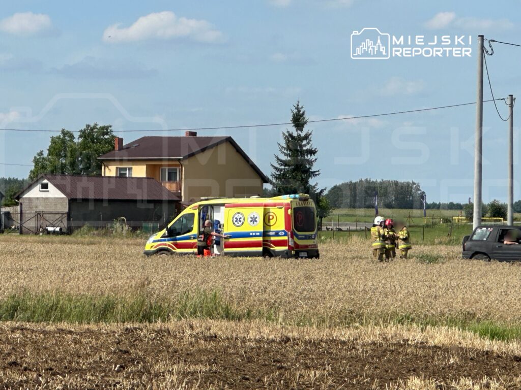 Zespół ratowniczy w mundurach rozmawia obok karetki na polu, w tle widoczny dom i drzewa.