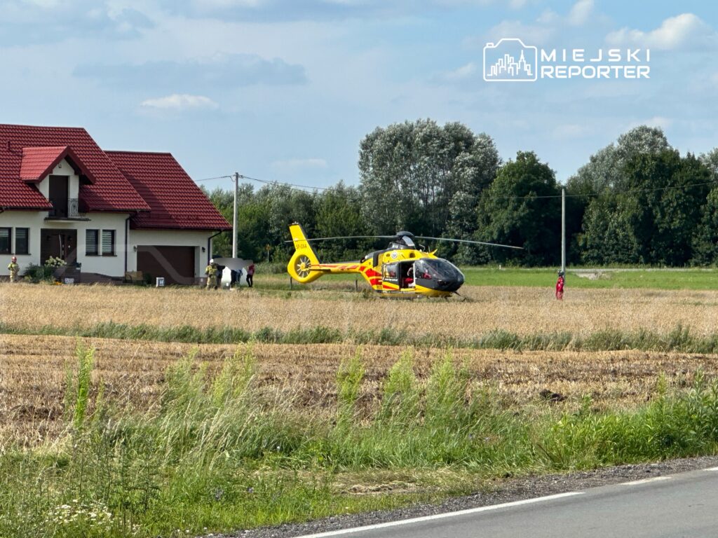 Śmigłowiec ratunkowy ląduje na polu obok domu, a ratownicy udzielają pomocy osobie w pobliżu.