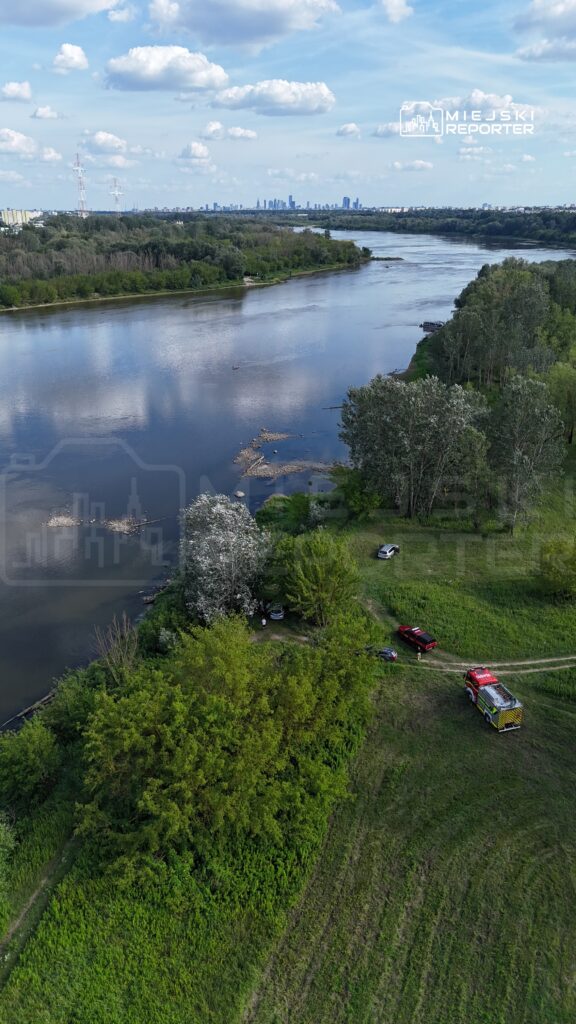 Widok na rzekę z zielonymi brzegami, w tle panorama miasta oraz samochód strażacki na łące.