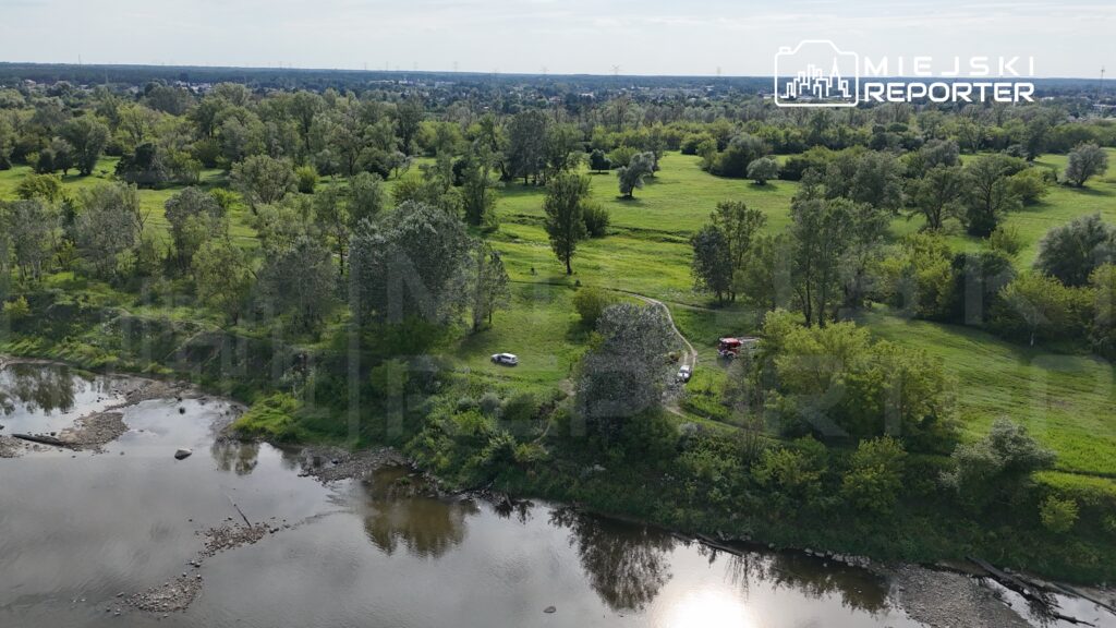 Na zielonym terenie nad rzeką stoją dwa samochody, a w oddali widać strażacki wóz.